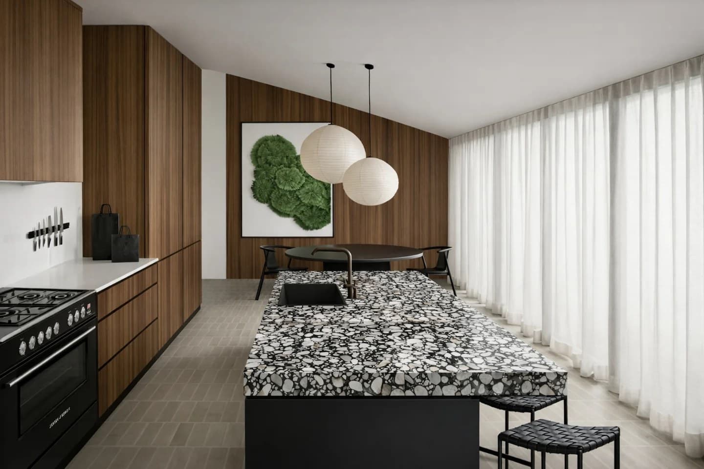 Kitchen in a 1970s home with walnut joinery, a bold stone island, twin paper pendants and floor-to-ceiling sheer curtains